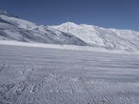 two people on a ski slope going down a snowy mountain side area in the foreground, with a tall snow covered hill behind them and a small lamp post in the top