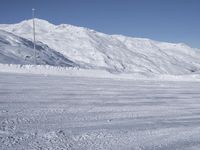 two people on a ski slope going down a snowy mountain side area in the foreground, with a tall snow covered hill behind them and a small lamp post in the top