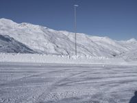 two people on a ski slope going down a snowy mountain side area in the foreground, with a tall snow covered hill behind them and a small lamp post in the top