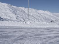 two people on a ski slope going down a snowy mountain side area in the foreground, with a tall snow covered hill behind them and a small lamp post in the top