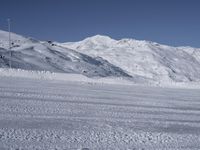 two people on a ski slope going down a snowy mountain side area in the foreground, with a tall snow covered hill behind them and a small lamp post in the top