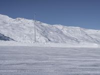 two people on a ski slope going down a snowy mountain side area in the foreground, with a tall snow covered hill behind them and a small lamp post in the top