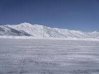 two people on a ski slope going down a snowy mountain side area in the foreground, with a tall snow covered hill behind them and a small lamp post in the top