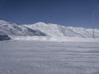 two people on a ski slope going down a snowy mountain side area in the foreground, with a tall snow covered hill behind them and a small lamp post in the top