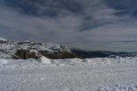 a person stands in snow with skis on in the mountains in the distance, a snow bank