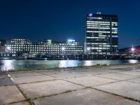 an empty bench sitting in front of the river at night in a city with high rise buildings