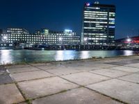 an empty bench sitting in front of the river at night in a city with high rise buildings