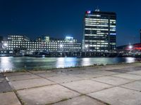 an empty bench sitting in front of the river at night in a city with high rise buildings