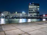 an empty bench sitting in front of the river at night in a city with high rise buildings