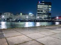 an empty bench sitting in front of the river at night in a city with high rise buildings