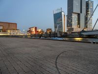 a park with benches next to water and buildings in the background at dusk and people standing next to it