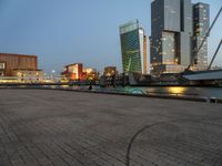 a park with benches next to water and buildings in the background at dusk and people standing next to it