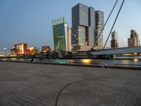 a park with benches next to water and buildings in the background at dusk and people standing next to it