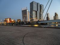 a park with benches next to water and buildings in the background at dusk and people standing next to it