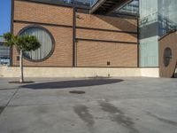 a parking area with a tree next to the entrance of a large building covered in brick walls