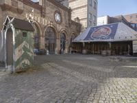 a small outdoor market with stalls in front of a brick building and a clock on the wall