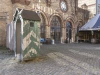 a small outdoor market with stalls in front of a brick building and a clock on the wall