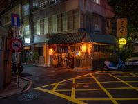 a small restaurant on a city street near parked cars at nighttime, illuminated by yellow lights