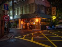 a small restaurant on a city street near parked cars at nighttime, illuminated by yellow lights