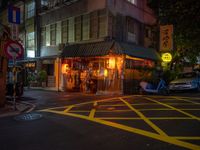 a small restaurant on a city street near parked cars at nighttime, illuminated by yellow lights