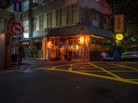 a small restaurant on a city street near parked cars at nighttime, illuminated by yellow lights