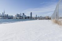 a fenced off area has snow covering the ground and is very snowy with a city in the background