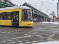 a yellow train moves down a wet street as people walk in the snow on either side of it