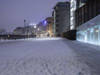 a snow covered walkway with buildings in the background at night time, along a waterfront