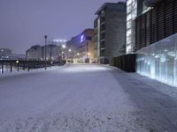 a snow covered walkway with buildings in the background at night time, along a waterfront