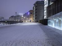 a snow covered walkway with buildings in the background at night time, along a waterfront