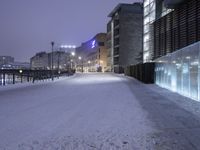 a snow covered walkway with buildings in the background at night time, along a waterfront