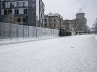 snow on the ground, building, and sidewalk next to it and fenced off with snow