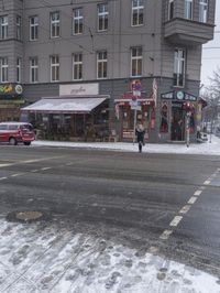 a city street full of stores covered with snow with people walking by them and cars parked on the side of it