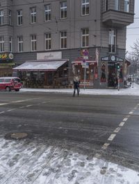 a city street full of stores covered with snow with people walking by them and cars parked on the side of it