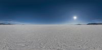 snow covered desert landscape with sun reflecting off clear blue sky above it and distant mountains in the distance