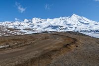 a mountain in the distance and dirt road to the right is an empty road with snow covered mountains in the background