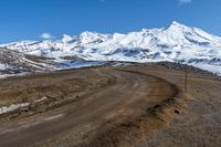 a mountain in the distance and dirt road to the right is an empty road with snow covered mountains in the background