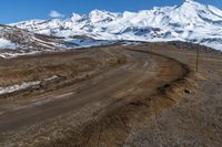 a mountain in the distance and dirt road to the right is an empty road with snow covered mountains in the background