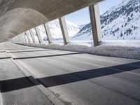 view from inside a curved bridge towards snow covered mountains in the distance with snow piles on either side