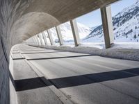 view from inside a curved bridge towards snow covered mountains in the distance with snow piles on either side