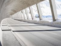 view from inside a curved bridge towards snow covered mountains in the distance with snow piles on either side