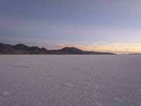 a snow covered plain surrounded by hills with the sun setting over the horizon, and tracks in the snow