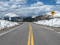 snow covers the roadway and snowy mountains on a sunny day, with a yellow warning sign in front of it