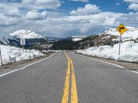 snow covers the roadway and snowy mountains on a sunny day, with a yellow warning sign in front of it