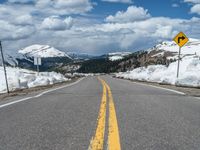 snow covers the roadway and snowy mountains on a sunny day, with a yellow warning sign in front of it