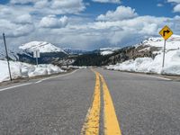 snow covers the roadway and snowy mountains on a sunny day, with a yellow warning sign in front of it