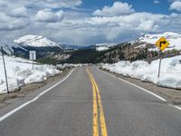 snow covers the roadway and snowy mountains on a sunny day, with a yellow warning sign in front of it