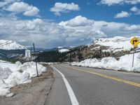 snow covers the roadway and snowy mountains on a sunny day, with a yellow warning sign in front of it