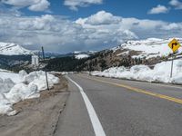 snow covers the roadway and snowy mountains on a sunny day, with a yellow warning sign in front of it