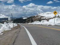 snow covers the roadway and snowy mountains on a sunny day, with a yellow warning sign in front of it